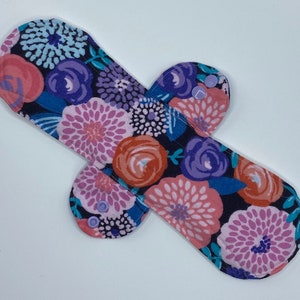 May include: A reusable cloth menstrual pad with a floral print in shades of pink, purple, blue, and red on a dark blue background. The pad has two wings with snaps for attaching to underwear.