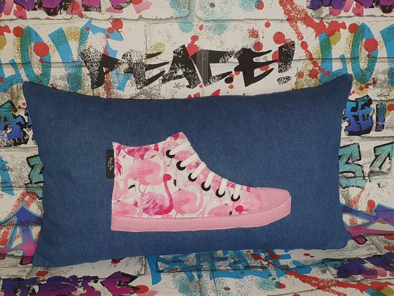 K&ouml;nnte beinhalten: Ein blaues Jeans-Kissen mit einem pinkfarbenen Flamingo-Druck-High-Top-Schuh-Design. Der Schuh ist pink mit schwarzen Schn&uuml;rsenkeln und einer wei&szlig;en Sohle.