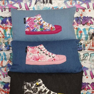 K&ouml;nnte beinhalten: Drei Kissenbez&uuml;ge aus Jeansstoff mit verschiedenen gemusterten High-Top-Sneakern, die darauf gestickt sind. Das erste Kissen hat einen pinkfarbenen Sneaker mit einem Blumenmuster, das zweite einen pinkfarbenen Sneaker mit einem Flamingomuster und das dritte einen schwarzen Sneaker mit einem tropischen Blatt-Muster.