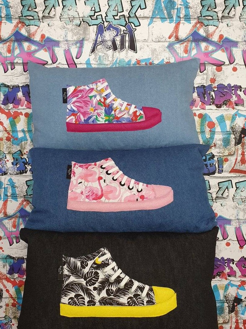 K&ouml;nnte beinhalten: Drei Kissenbez&uuml;ge aus Jeansstoff mit verschiedenen gemusterten High-Top-Sneakern, die darauf gestickt sind. Das erste Kissen hat einen pinkfarbenen Sneaker mit einem Blumenmuster, das zweite einen pinkfarbenen Sneaker mit einem Flamingomuster und das dritte einen schwarzen Sneaker mit einem tropischen Blatt-Muster.