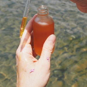 May include: A clear glass bottle with a dropper filled with a golden liquid. The bottle is being held by a hand over a body of water.
