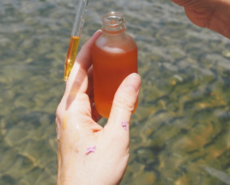 May include: A clear glass bottle with a dropper filled with a golden liquid. The bottle is being held by a hand over a body of water.