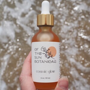 May include: A glass bottle with a gold lid and dropper, filled with a light orange liquid. The label reads "Of The Sun Botanicals" and "Cosmic Glow" with a small illustration of a sun and flowers. The bottle is labeled "2 oz / 60 ml".