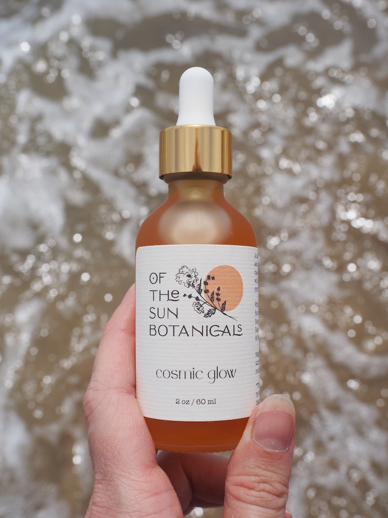 May include: A glass bottle with a gold lid and dropper, filled with a light orange liquid. The label reads "Of The Sun Botanicals" and "Cosmic Glow" with a small illustration of a sun and flowers. The bottle is labeled "2 oz / 60 ml".