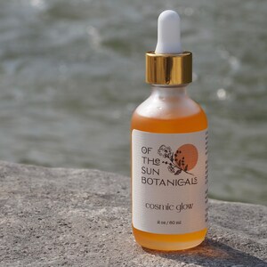 May include: A clear glass bottle with a gold lid and a label that reads "Of The Sun Botanicals" and "Cosmic Glow". The bottle contains a yellow liquid and is sitting on a gray rock.