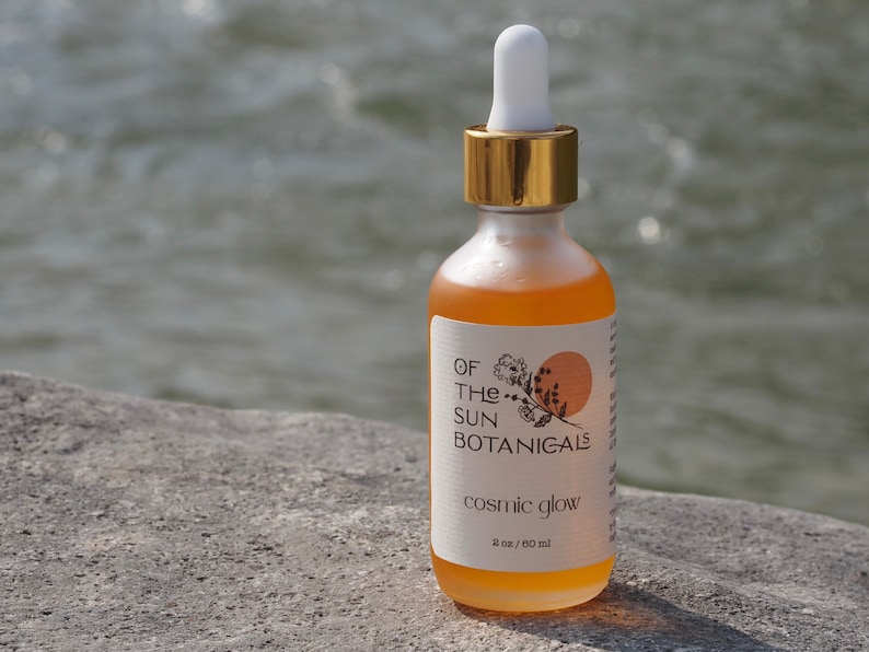 May include: A clear glass bottle with a gold lid and a label that reads "Of The Sun Botanicals" and "Cosmic Glow". The bottle contains a yellow liquid and is sitting on a gray rock.
