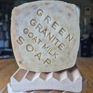 May include: A bar of goat milk soap with the words "Green Granite Soap" embossed on the top. The soap is sitting on a wooden soap dish.