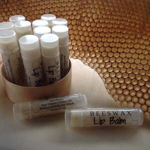 May include: A close-up of a group of lip balm tubes in a cardboard container. The lip balms are labeled "Beeswax Lip Balm" and have a list of ingredients including raw organic coconut oil, beeswax from the Fox Islands, and vitamin E oil. The lip balms are arranged in a circle around the container. The container is sitting on a white surface. A honeycomb pattern is visible in the background.