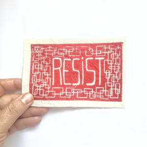 May include: A rectangular art print with the word "RESIST" in bold white letters on a red background. The background features a white geometric pattern. The print is held by a hand against a white backdrop.