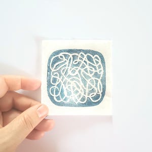 May include: A small, square artwork featuring a blue background with a white, swirling line design. The artwork is held by a hand against a white background. The design is abstract and appears to be hand-painted.