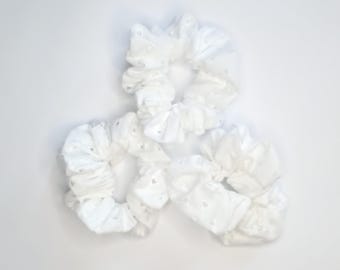 May include: Three white eyelet fabric scrunchies arranged on a white background. The scrunchies are made of a soft, textured material with small, decorative cut-out patterns. These hair accessories are designed to hold hair in place.