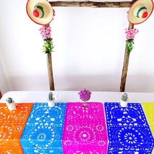 May include: Five colorful paper table runners with intricate cut-out designs. The runners are orange, blue, pink, blue, and yellow. The runners are laid out on a white table with small potted plants on either side.