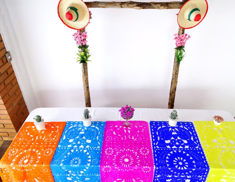 May include: Five colorful paper table runners with intricate cut-out designs. The runners are orange, blue, pink, blue, and yellow. The runners are laid out on a white table with small potted plants on either side.