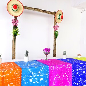 May include: A table setting with colorful table runners featuring a traditional Mexican paper cutting design. The table runners are orange, blue, pink, and yellow. There are small potted cacti and a vase of flowers on the table. Two straw hats with red and green trim hang from a wooden frame above the table.