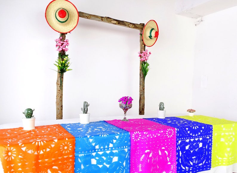 May include: A table setting with colorful table runners featuring a traditional Mexican paper cutting design. The table runners are orange, blue, pink, and yellow. There are small potted cacti and a vase of flowers on the table. Two straw hats with red and green trim hang from a wooden frame above the table.