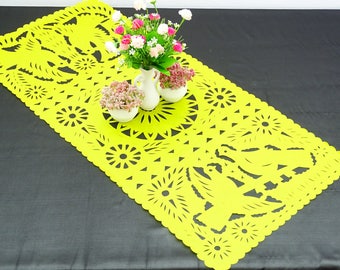 May include: A yellow paper table runner with a delicate cut-out design featuring flowers and birds. The runner is decorated with a scalloped edge and is displayed on a black surface with two small white vases filled with pink and white flowers.