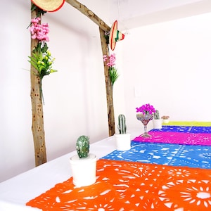 May include: A table setting with a white tablecloth and colorful paper table runners. The table runners are decorated with a traditional Mexican design. There are small potted cacti and a glass of water on the table.