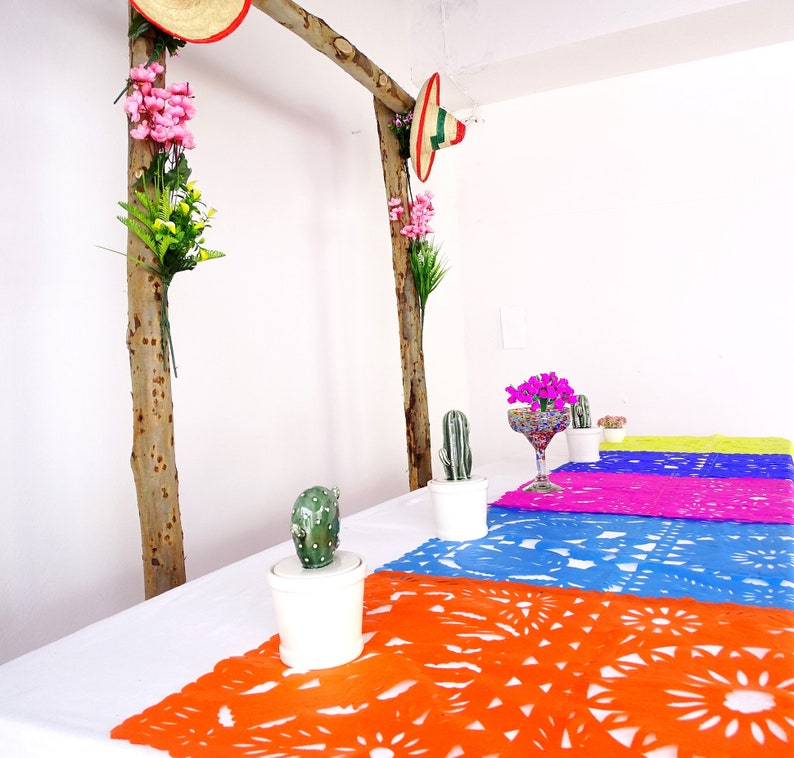 May include: A table setting with a white tablecloth and colorful paper table runners. The table runners are decorated with a traditional Mexican design. There are small potted cacti and a glass of water on the table.