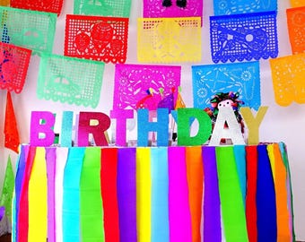 May include: A table decorated with a vibrant, multi-colored vertical paper fringe skirt and large, glittery, multi-colored letters spelling out the word BIRTHDAY. The background features several colorful, intricate, die-cut paper banners.
