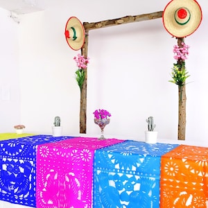 May include: A table setting with a colorful paper table runner featuring a traditional Mexican design. The table runner is made of bright yellow, blue, pink, and orange paper. The table is decorated with small potted cacti and flowers.