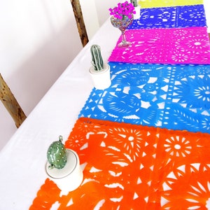 May include: A table setting with five colorful paper table runners with intricate cut-out designs. The runners are in shades of orange, blue, pink, purple, and yellow. Each runner is placed on a white tablecloth. Small potted cacti and a glass vase with pink flowers are also on the table.