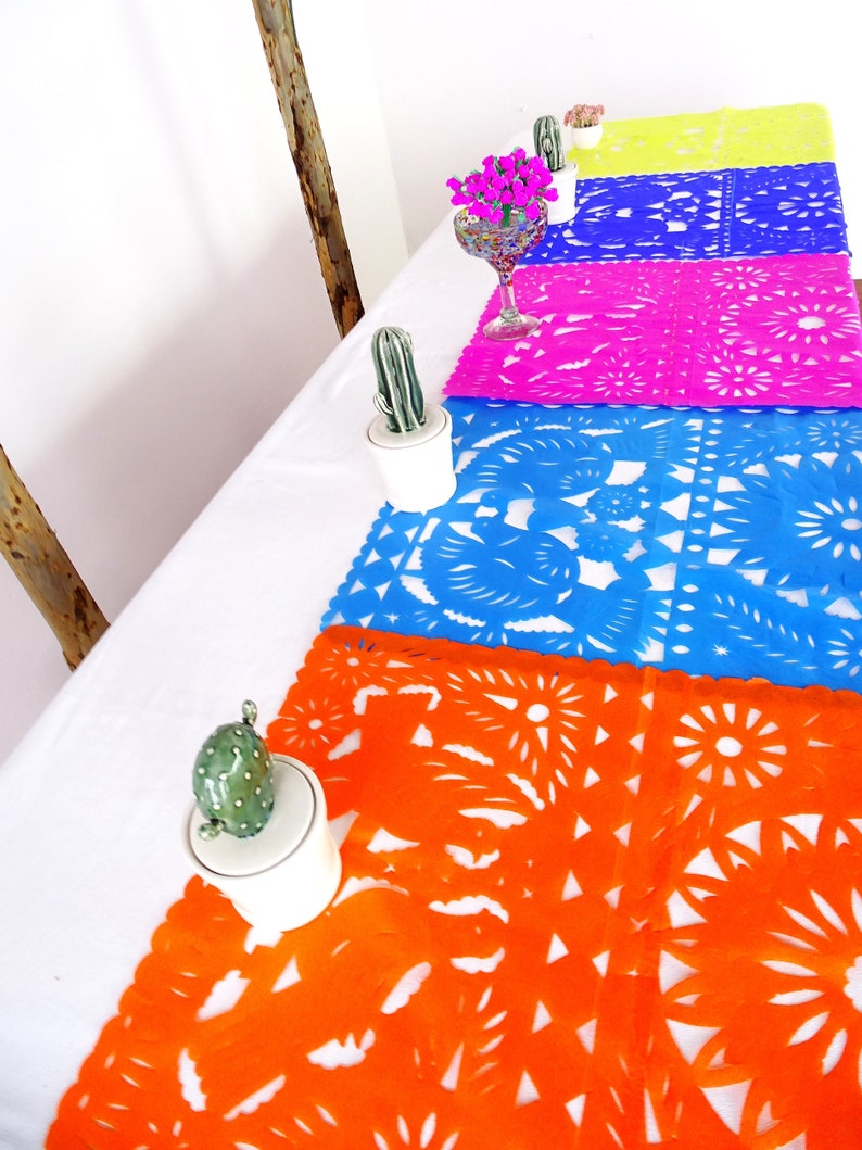 May include: A table setting with five colorful paper table runners with intricate cut-out designs. The runners are in shades of orange, blue, pink, purple, and yellow. Each runner is placed on a white tablecloth. Small potted cacti and a glass vase with pink flowers are also on the table.