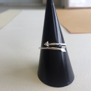 Könnte beinhalten: Ein silberner Ring mit zwei Pfeilmotiven. Der Ring wird auf einem schwarzen, kegelförmigen Ringständer präsentiert.