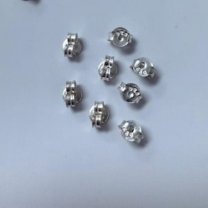 Puede incluir: Una colección de cierres de pendientes plateados, dispersos sobre una superficie gris claro. Los cierres de pendientes tienen forma de mariposa, diseñados para asegurar los pendientes. La imagen es una vista superior, mostrando los detalles de los hallazgos de joyería.