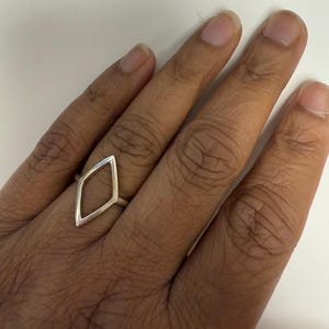 May include: A silver ring featuring a diamond-shaped design. The ring is worn on a hand, with the skin tone and nail detail visible. The ring's minimalist style is set against a neutral background.