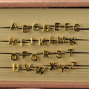 May include: Gold alphabet letters are arranged in order on a closed book with a pink cover. The letters are slightly raised from the surface of the book.