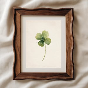 May include: A framed watercolour print of a four-leaf clover. The artwork features a green clover with a delicate stem against a cream-coloured background. The frame is made of dark brown wood with a wavy edge.