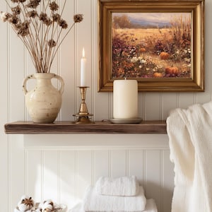 May include: A rustic wooden shelf displays a cream-colored vase with dried flowers, a lit candle in a brass holder, and a large pillar candle. A framed painting of a field with pumpkins hangs above. White towels are stacked below.