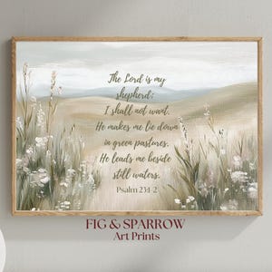 Op de afbeelding: Een ingelijste kunstdruk met een pastorale landschapsscène en tekst uit Psalm 23:1-2. Het kunstwerk toont een veld met hoog gras en wilde bloemen in gedempte groen-, beige- en grijstinten. De tekst luidt "De Heer is mijn herder..." en de merknaam "FIG & SPARROW Art Prints" staat onderaan.