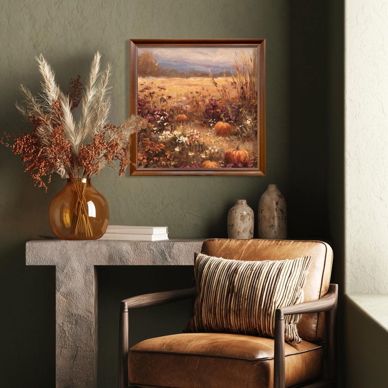 May include: A framed landscape painting depicts a field with pumpkins, flowers, and tall grass. A brown vase with dried flowers sits on a stone table. A leather chair with a striped pillow is in the foreground.