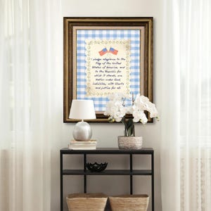 May include: Framed artwork featuring the Pledge of Allegiance with two American flags. The artwork has a gold frame, a blue and white gingham background, and star-shaped embellishments. A white lamp and flowers are on a black table.