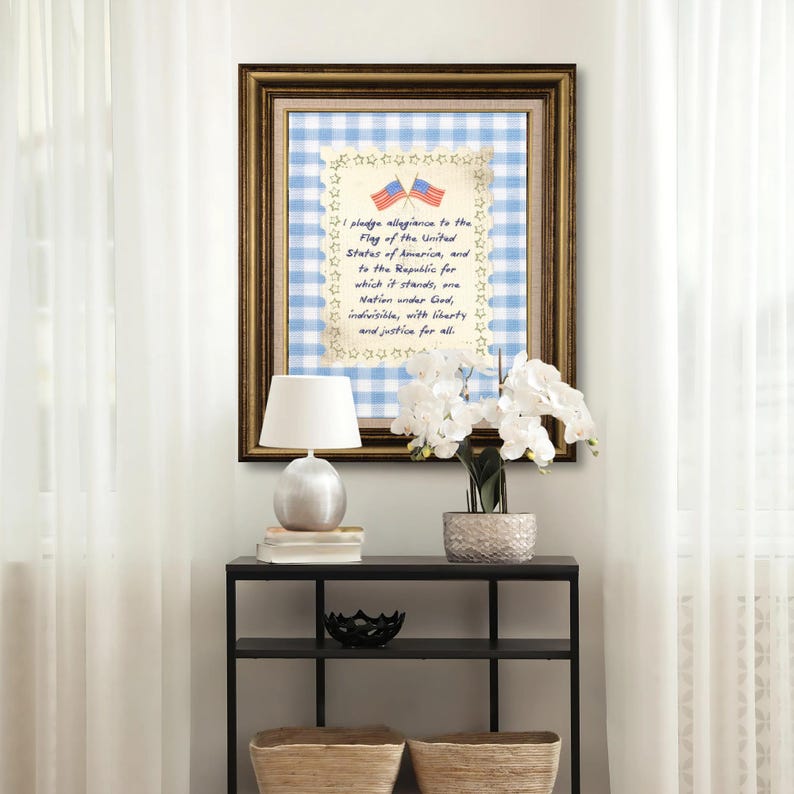 May include: Framed artwork featuring the Pledge of Allegiance with two American flags. The artwork has a gold frame, a blue and white gingham background, and star-shaped embellishments. A white lamp and flowers are on a black table.