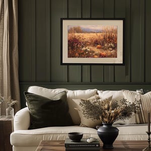 May include: A framed landscape painting depicting a field of wildflowers and pumpkins in autumn colors hangs above a white sofa with decorative pillows. A black vase with dried flowers sits on a wooden coffee table.