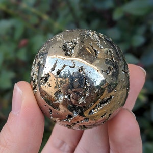 Könnte beinhalten: Eine polierte Pyritkugel, auch bekannt als Narrengold, wird in einer Hand gehalten. Die Kugel hat eine metallisch goldene Farbe mit dunklen, strukturierten Einschlüssen. Der Hintergrund ist verschwommenes grünes Laub. Durchmesser ca. 6 cm.