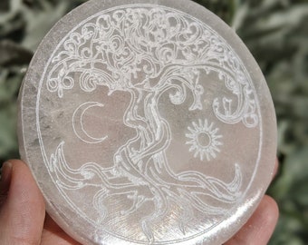 May include: A round, translucent, selenite coaster with an etched design of a tree, crescent moon, and sun. The coaster is held in a hand, with a blurred green background. The tree design is centered, with the moon and sun on either side.