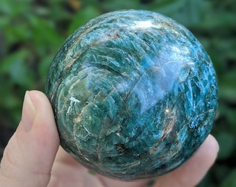 May include: A polished, spherical gemstone in shades of teal and green, held in a hand. The stone's surface has a textured, crystalline appearance, with variations in color and light reflection. The background is a blurred green.