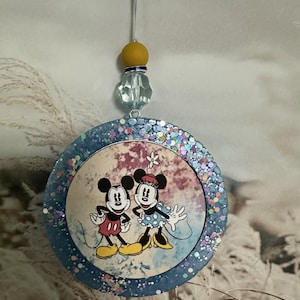 May include: A round ornament featuring Mickey and Minnie Mouse. The image of the characters is set against a watercolor-style background. The ornament has a blue glitter border and is suspended by a white string with a yellow bead and a clear, faceted bead.