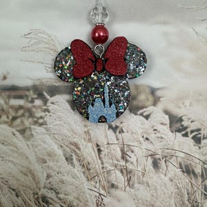 May include: A glittery Minnie Mouse-shaped ornament with a red bow and a blue castle design. The ornament is suspended by a clear string with a red bead and a clear crystal bead. The background features tall, dried grasses.