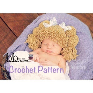 May include: A crochet pattern for a baby hat with pigtails. The hat is made of yarn in a light brown color and has two white bows on top. The hat is being worn by a baby sleeping in a purple blanket.