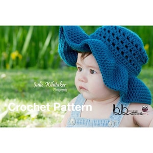 May include: A baby wearing a blue crocheted sun hat with a wide brim. The hat has a lacy pattern and is sitting on the baby's head. The baby is looking off to the side and has a serious expression on their face. The background is a blurry green field.