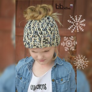 May include: A girl wearing a blue denim jacket over a white t-shirt with the word "unicorns" printed on it. She is wearing a crocheted beanie with a messy bun on top. The beanie is a mix of white, gray, and black yarn.