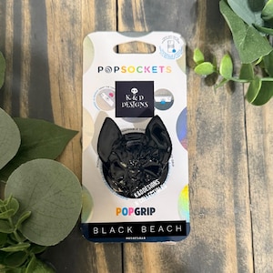 May include: A black PopSockets PopGrip phone accessory featuring a sculpted cat head design. The product is mounted on its original retail packaging, which displays the brand name PopSockets, the K&D Designs logo, and the text Black Beach at the bottom.