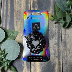 May include: A black PopSocket with a crystal heart design, framed by a decorative border and butterfly accents. The PopSocket is attached to a holographic backing card with the text "BLACK FRAME CRYSTAL".