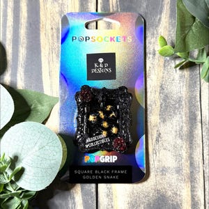 May include: A PopGrip phone accessory on a holographic backing card. The accessory features a square black frame with glitter, containing a coiled golden snake and two small red roses. Text on the card includes PopSockets, K&D Designs, and Collectibles.