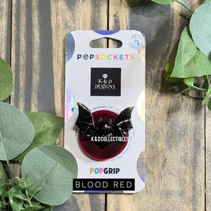 May include: A PopSockets PopGrip phone accessory featuring a black bat-shaped swappable top over a circular blood red base. The product is mounted on a white retail card with the text PopSockets, K&D Designs, K&DCollectibles, and Blood Red.