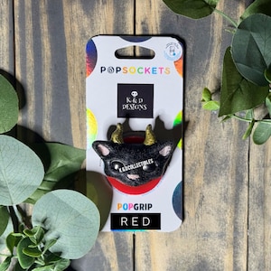 May include: A black cat-shaped phone grip with small gold horns and large black eyes, mounted on a white backing card labeled PopSockets, K&D Designs, and K&DCollectibles. The base of the phone grip is red, and the card features the word RED.